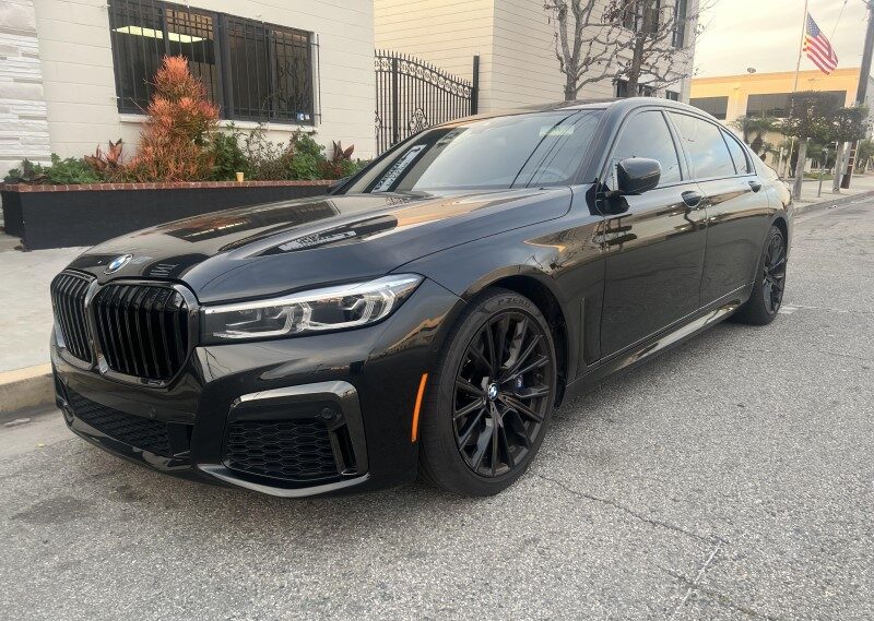 A black BMW sedan is parked on a street in front of a building with a flagpole displaying the American flag. The car's sleek auto body, complemented by black wheels and shining headlights, exudes sophistication.
