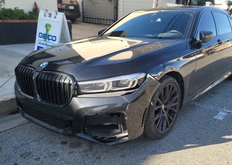 A black BMW with minor front bumper damage is parked on the street. A sign in the background reads "GEICO Auto Body Repair.