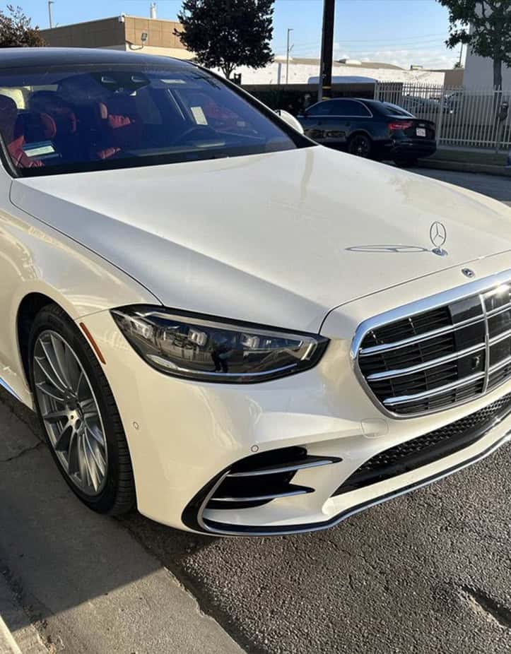 A white Mercedes-Benz car is parked on the side of the road in front of a fence and a smaller black car, near a Collision Center. The background includes trees and a building.