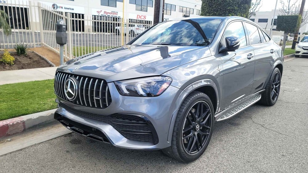 A silver Mercedes-Benz SUV, recently undergoing expert collision repair, is parked on a street in front of a fence and buildings. The vehicle boasts a prominent front grille and black alloy wheels.