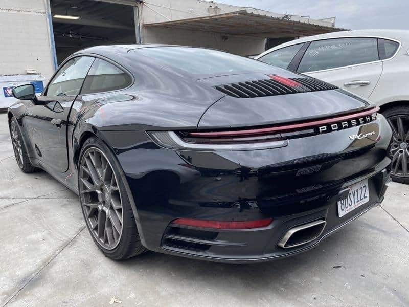A black Porsche 911 Carrera is parked next to a white vehicle in front of an industrial building with an open garage door, a notable facility known for its top-tier collision repair services.