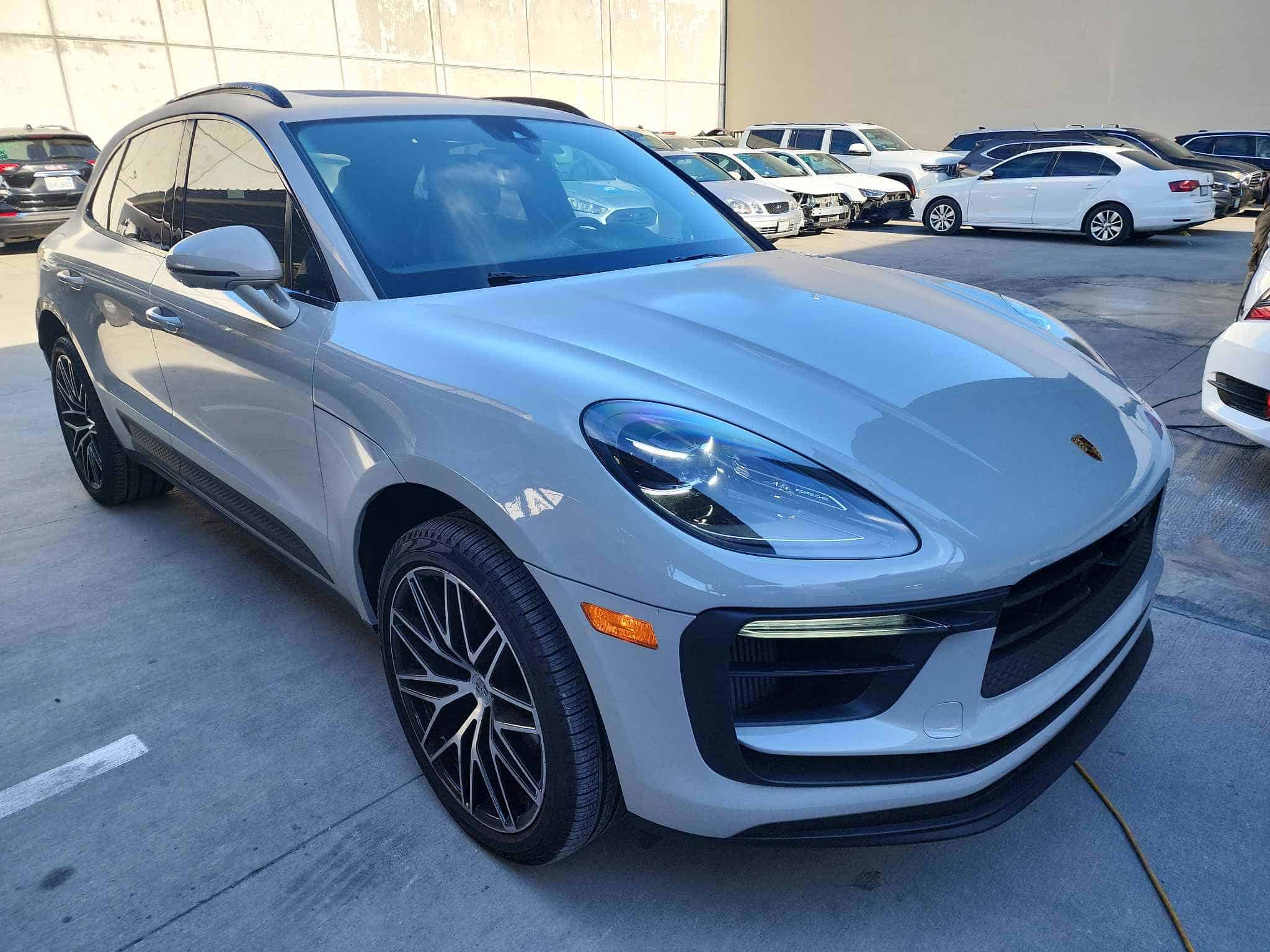 A silver porsche SUV, recently back from auto body repair, is parked in an outdoor lot with several other vehicles. The sun is shining, creating reflections on the car's surface.