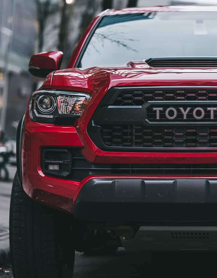 Close-up of the front of a certified red Toyota pickup truck parked on a city street. The headlights and grille are prominently visible, showcasing the impeccable work done at Toyota Collision Repair.