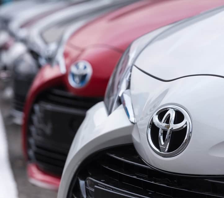 Close-up of the grilles and front bumpers of several parked Toyota cars in a row, showcasing Toyota logos and different colors including red and silver, at a certified body shop.